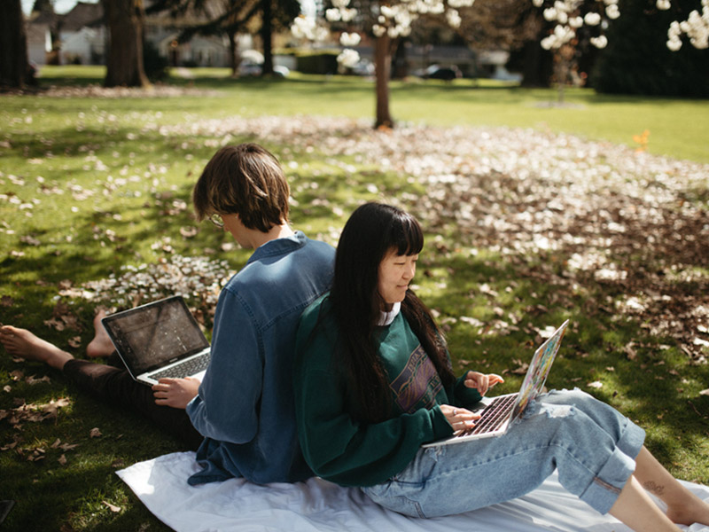 Two students sit back to back on a blanket on the Great Lawn working on their laptops with fallen leaves behind them. 