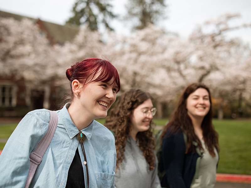 Three students smile while walking past cherry blossom trees.