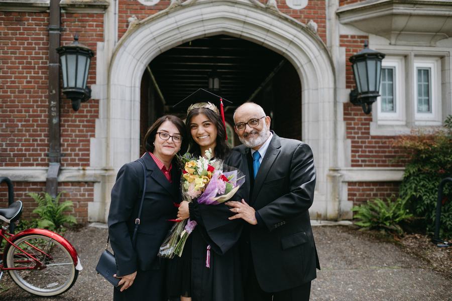 A family of three is standing next to each other in front of an academic building and one is dressed in a graduation robe and cap.