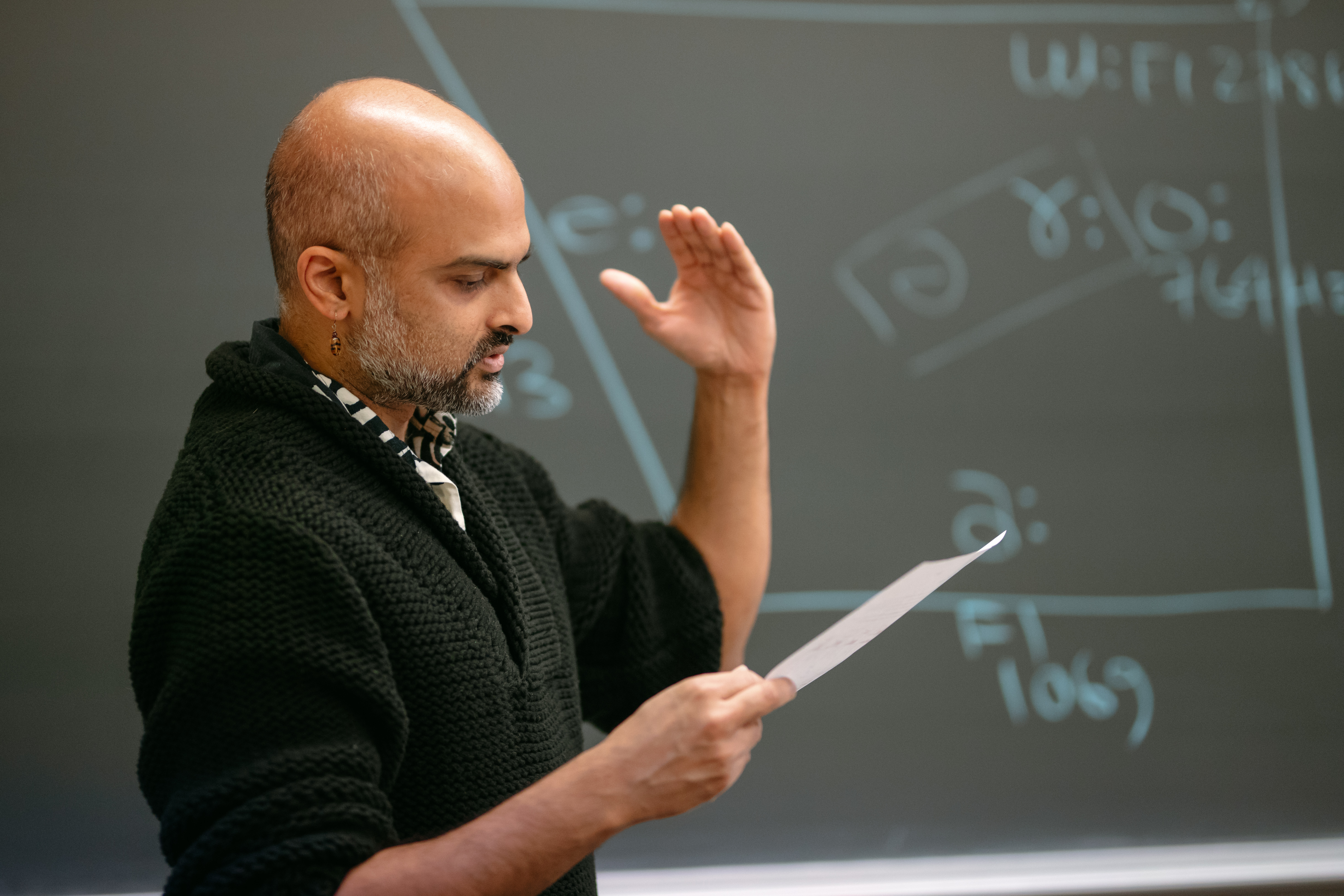 A professor standing in front of a chalkboard, reading from a paper.