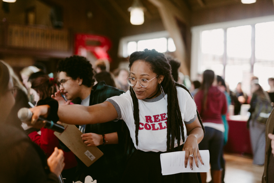 A student in a white and red Reed College shirt holds up a microphone to someone out of shot while many people crowd the background.