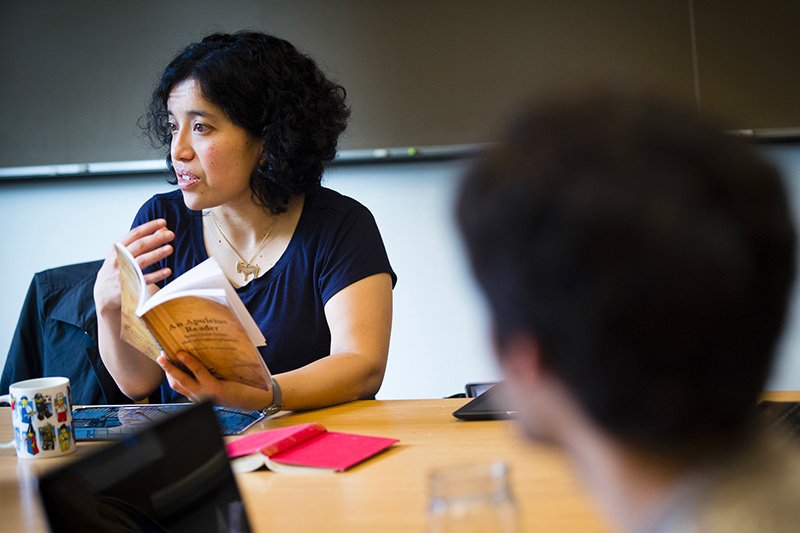 A professor holds a book open in front of them and explains something in the classroom with a student in the foreground. 