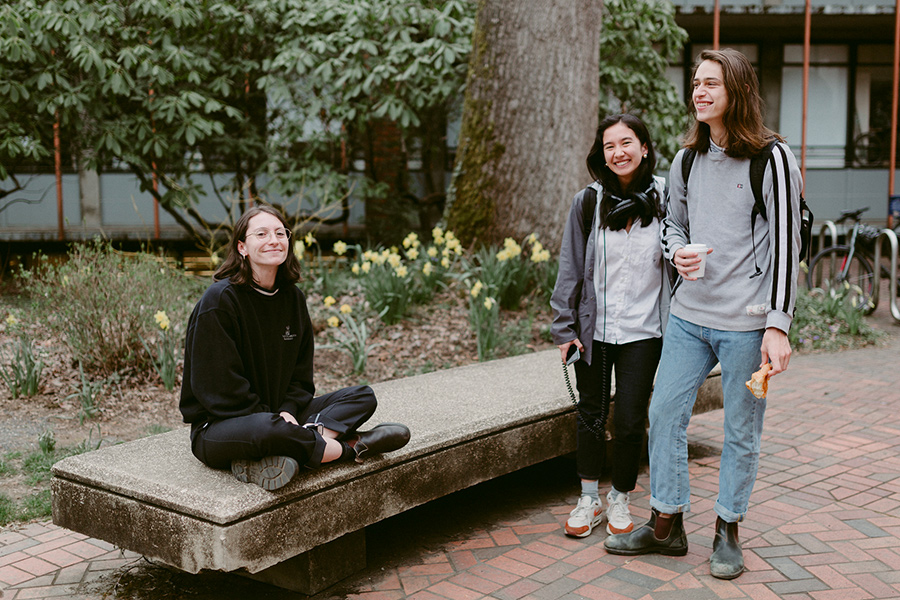 Three students surround a bench near the library, two of which are standing together and the other of which is sitting and smiling at the camera. 