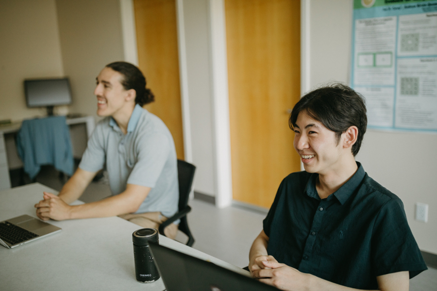 Two students sit smiling in class next to each other while looking at something out of shot.