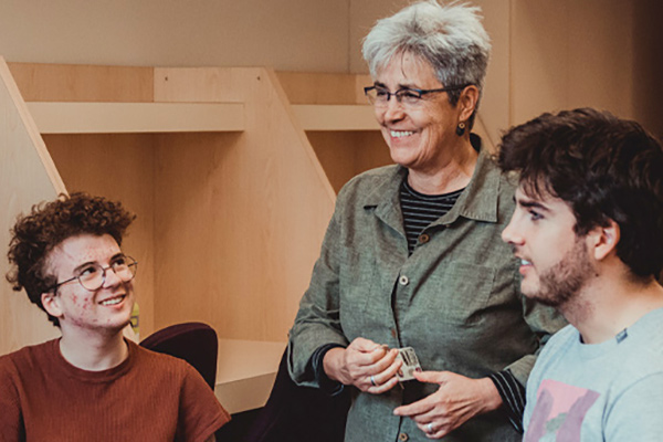 A professor stands behind two seated students talking to them about something out of shot.