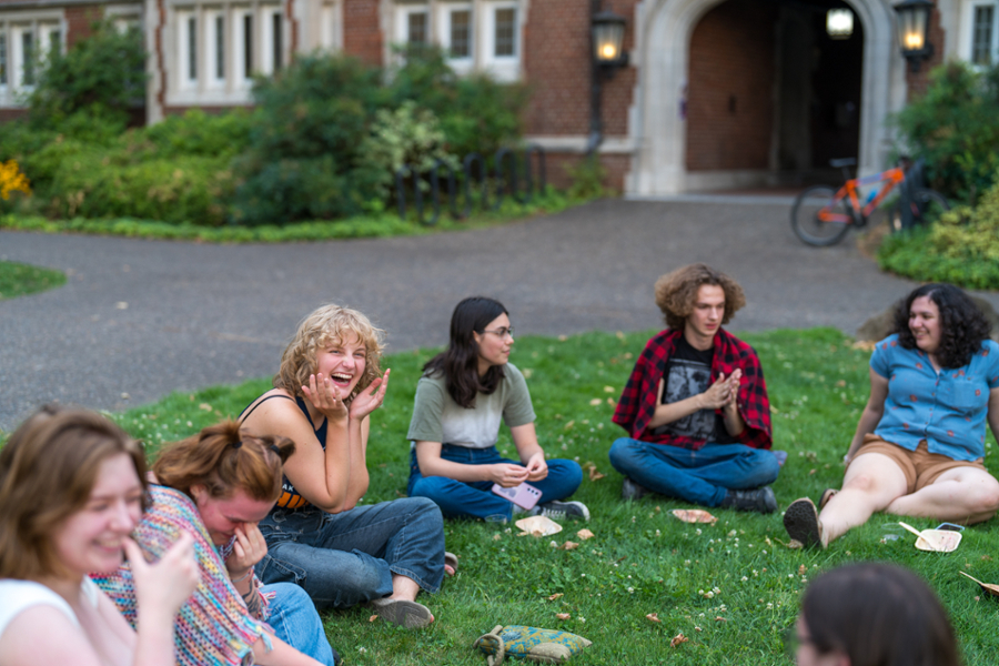 A group of students sits laughing in a circle in the grass with the Sally Port in the background.