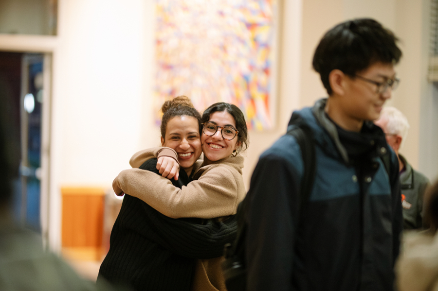 Two students hug and smile at the camera while another student walks by in the foreground.