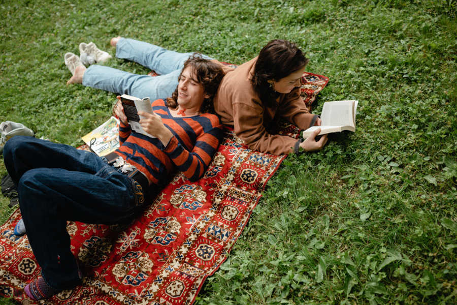 Two students lay on a rug in the grass reading books, one on their stomach and the other on their back with their head leaning against their friend.