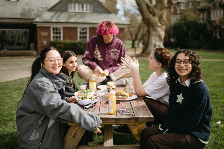 A group of five students sits and eats lunch at a picnic bench in the quad, one of which is sat on the table top.