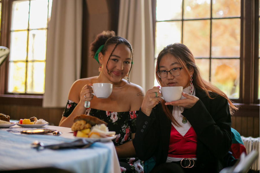 Two students sit in the Student Union and hold up cups of tea with plates piled with food in front of them. 