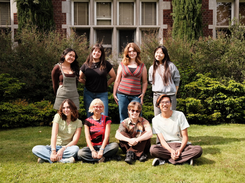 A group of students, four standing and four sitting, are smiling in front of a red brick building.