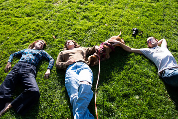 Three students lay on their backs in the grass, two of whom are petting a dog laying between them. 