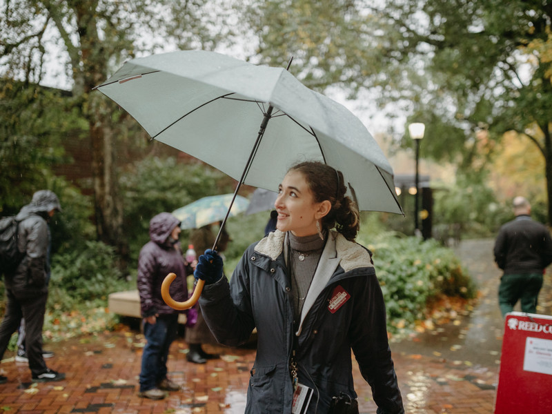 A admission tour guide is leading a tour on a rainy day while holding an umbrella and looking up to the sky.