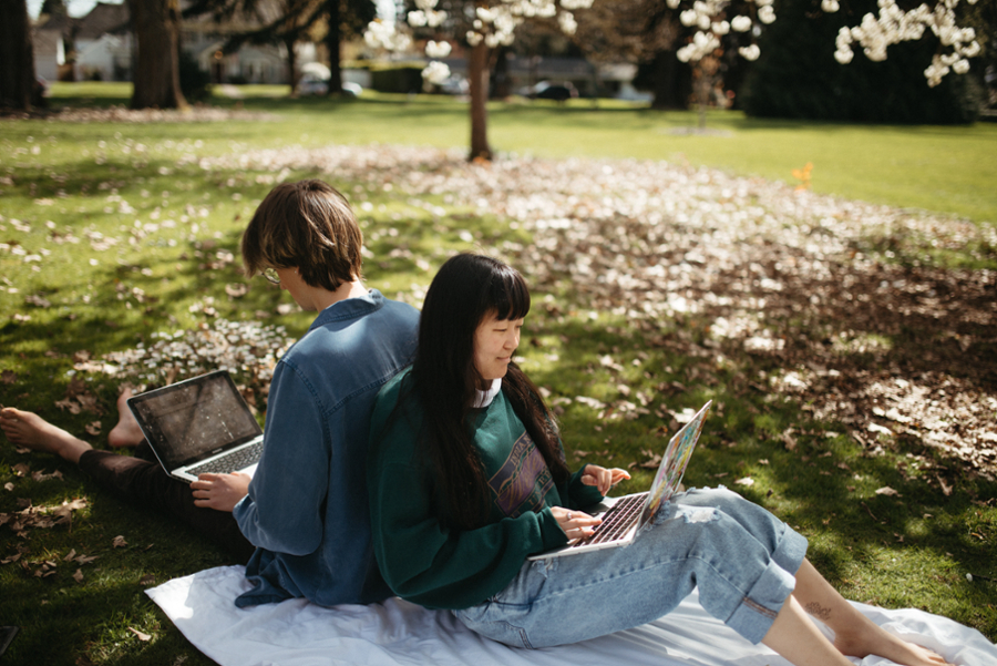 Two students sitting back to back in the grass and working on their laptops