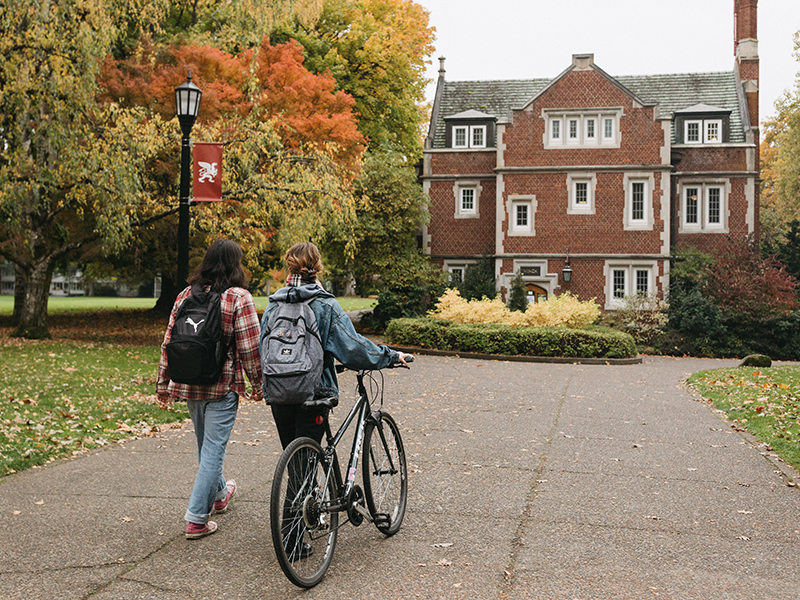 Two students walk toward Old Dorm Block with a bicycle.