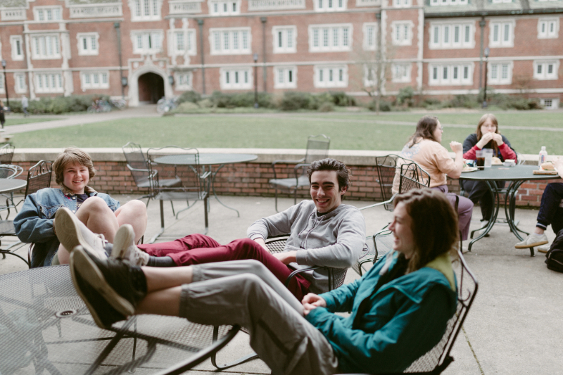 A group of students are sitting at a table outside, laughing and eating together.
