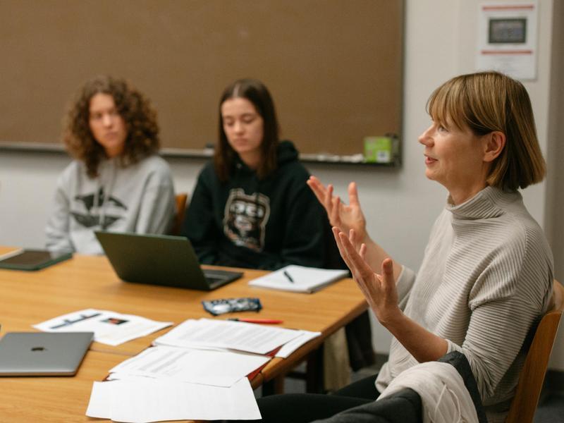 A professor gestures with her hands while sitting with students at a table covered with laptops, notebooks, and paper.