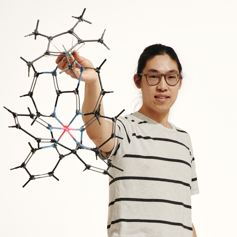 A portrait photo of Reed College student Joshua Tsang. He is holding up a 3-D model of a complex molecule