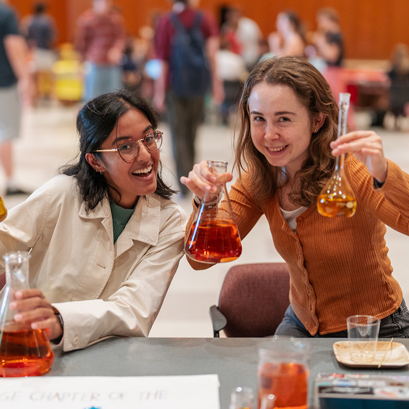 Two students sit at a table at an activity fair holding beakers up and smiling