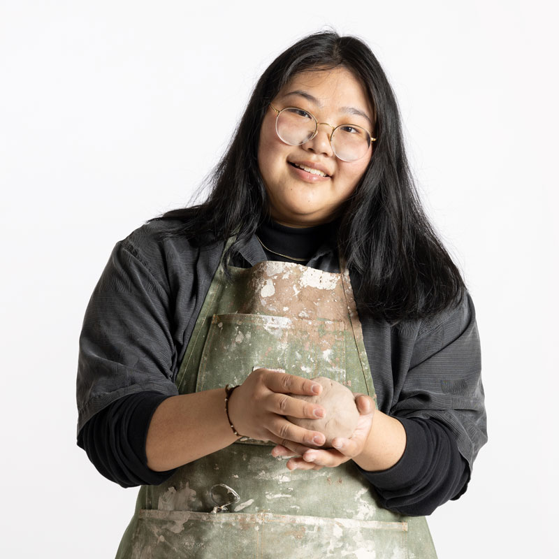 A portrait photo of Stephanie Shu, who is holding a ball of clay and is wearing a green apron