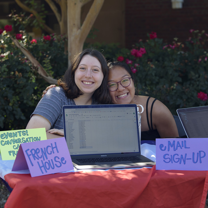 Two students sit at a table outside. They are smiling, and signs on the table say French House, Email sign-up, and Events, Conversation.