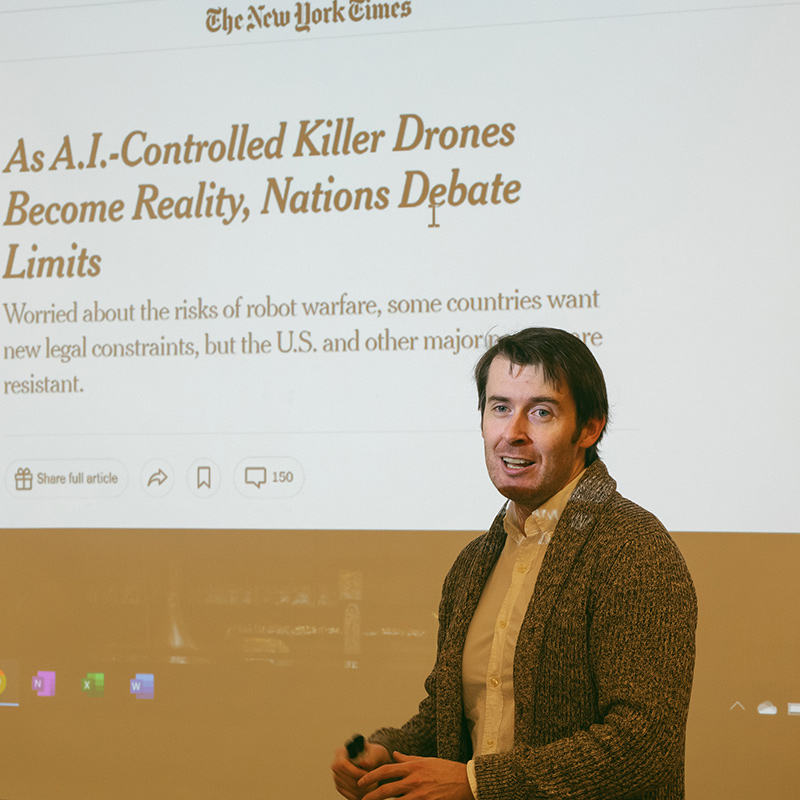 Reed German Professor Jake Fraser stands in a classroom in front of a slide that shows an article from the New York Times about robot warfare.
