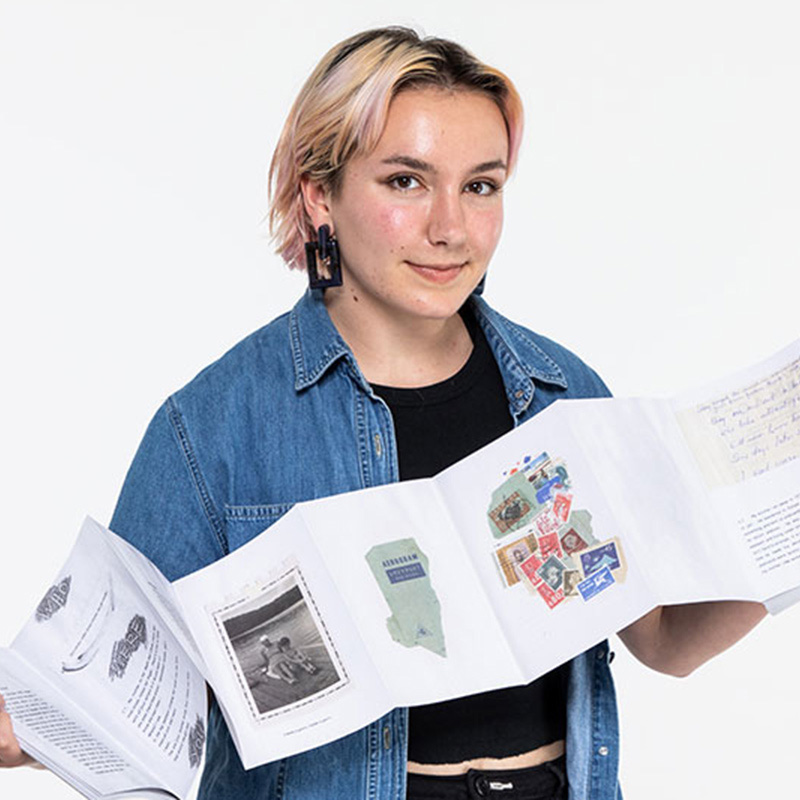 Photo portrait of student Emma McNeel. She is unfolding an artist's book, which shows a black and white photo, text, and a collage of stamps.