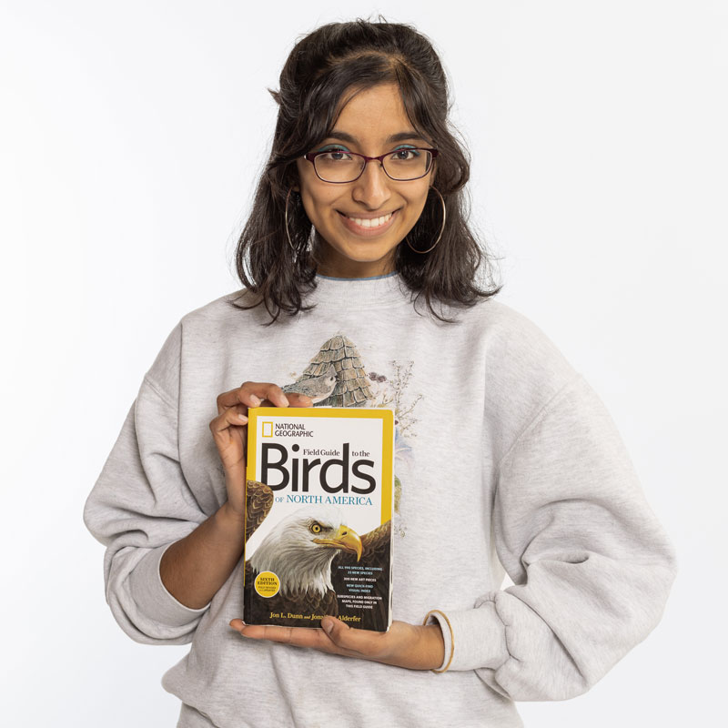A portrait photo of Ananke Garani Krishnan, who is holding up the book "FIeld Guide to the Birds of North America"