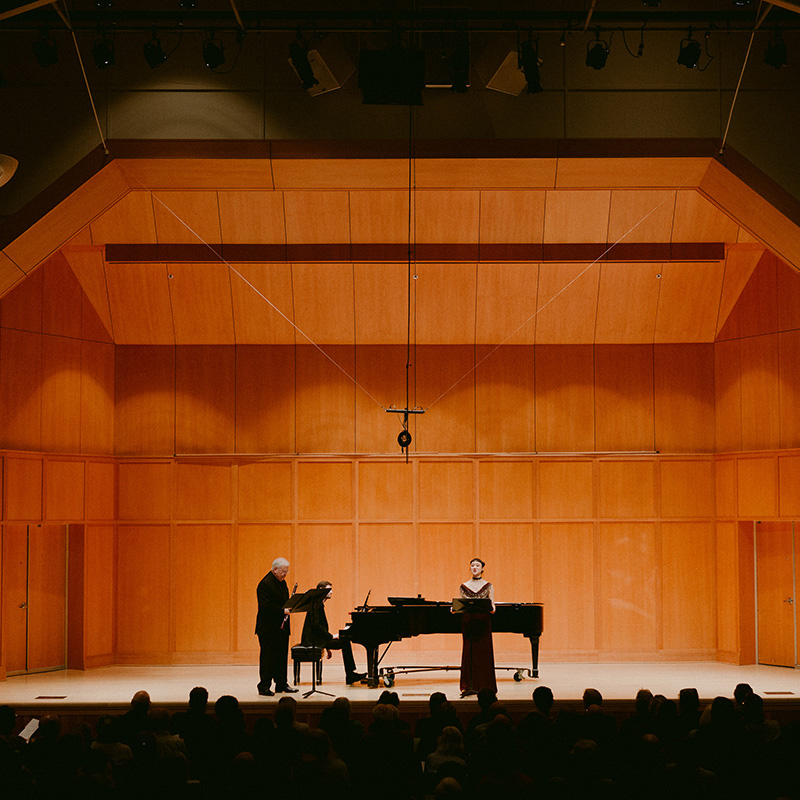 Three figures on the Kaul Auditorium stage, one seated at the piano, one holding a wind instrument, and a third talking at a podium.