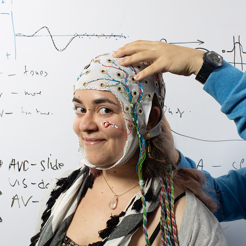 A Reed student getting an electro-encephalography or EEG cap adjusted on her head in front of a whiteboard filled with calculations. 