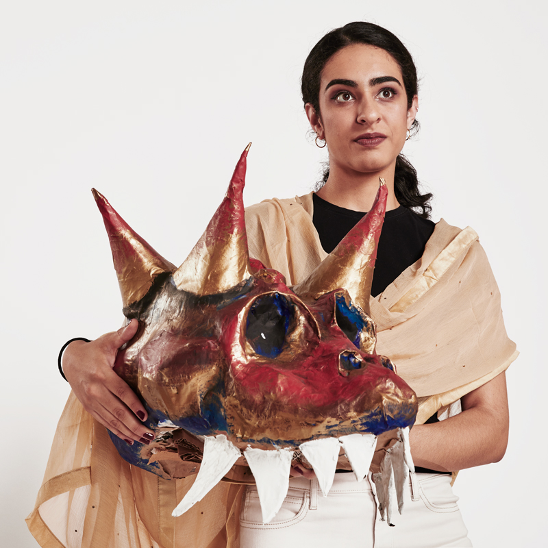 A portrait photo of Aziza Afzal, who is holding a theatre prop of a dragon's head