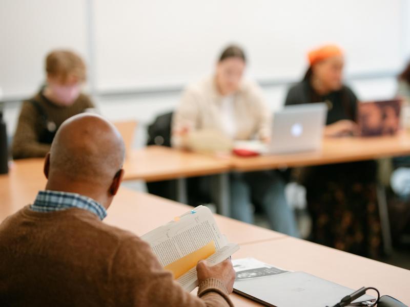 An over-the-shoulder view of a professor holding an open book while addressing a classroom of students.