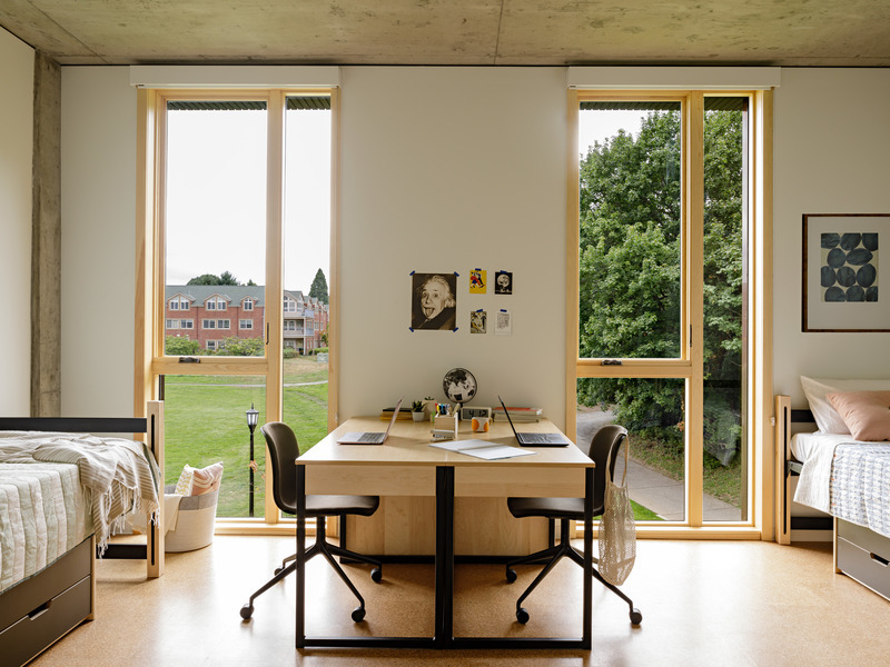 Interior of a dorm room with a shared desk, two beds, and two windows overlooking campus.