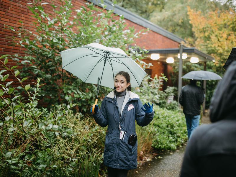 A student holds an umbrella and gestures with their hands while giving a campus tour outside.