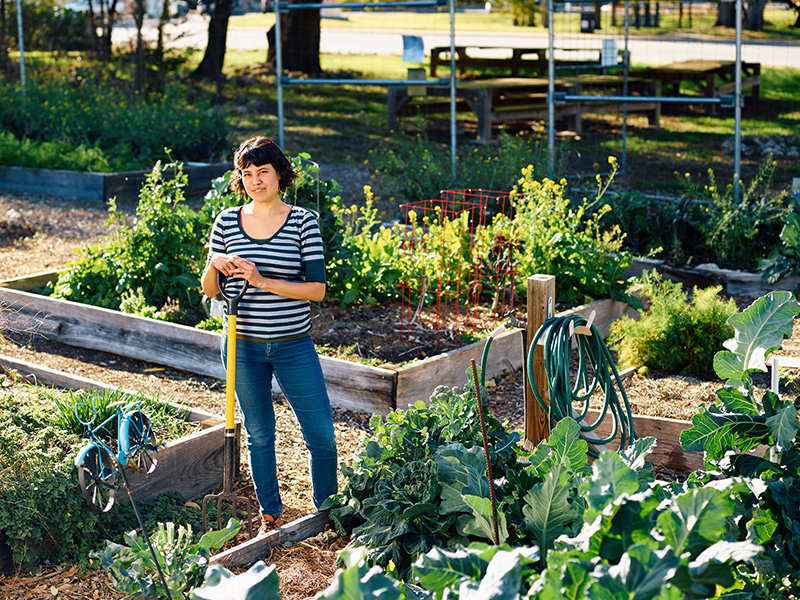 Sari Albornoz, a Reed College graduate, stands outside in a garden, smiling while holding a garden fork