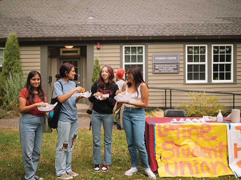 Four Reed College students stand beside a table with a poster that says "Latine Student Union" while eating Latin food