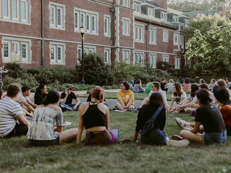 A group of about 20 Reed students sit around a circle on a green lawn during an orientation event