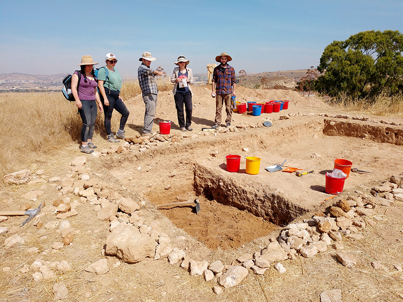 Men and women gather around and talk around a desert trench with rocks, digging tools, and buckets surrounding it