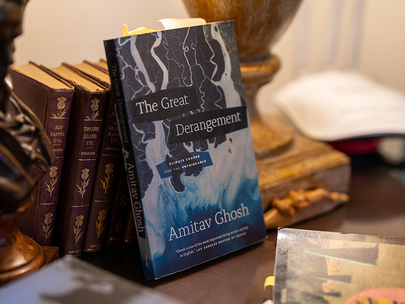 A copy of a book titled "The Great Derangement" by Amitav Ghosh propped up against a short stack of other books on a table.