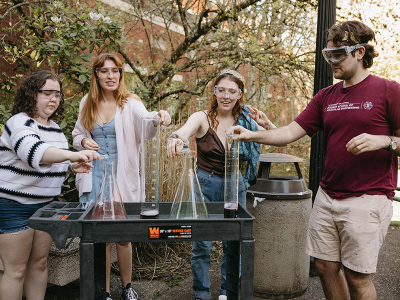 Four Reed College students wearing safety glasses stand outside around a cart, pouring chemicals in four large test tubes.
