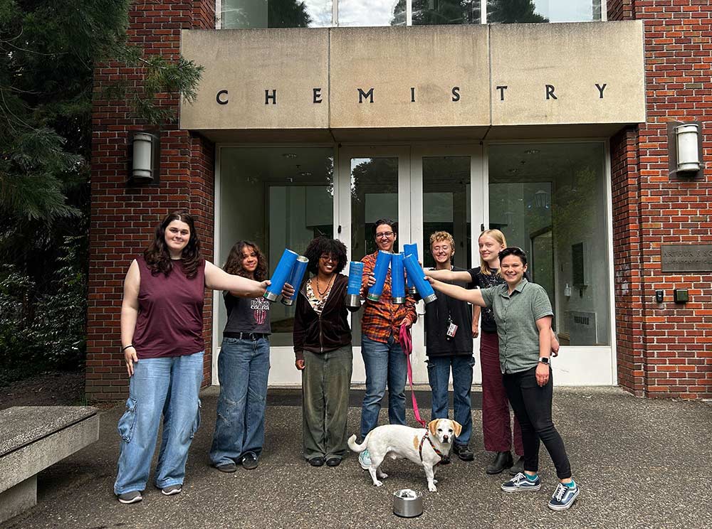 Six students, along with their professor and a cute dog, posing outside for a group photo
