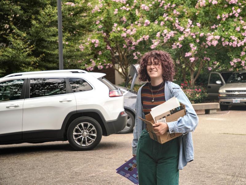 Student carrying a cardboard box in a parking lot.