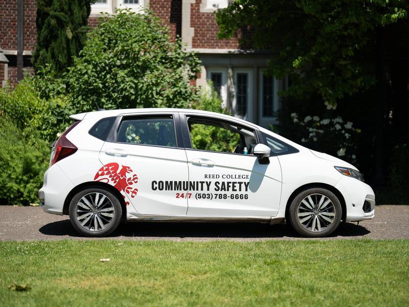 A white car with a Reed College Community Safety decal on the side parked outside of a brick building on campus.