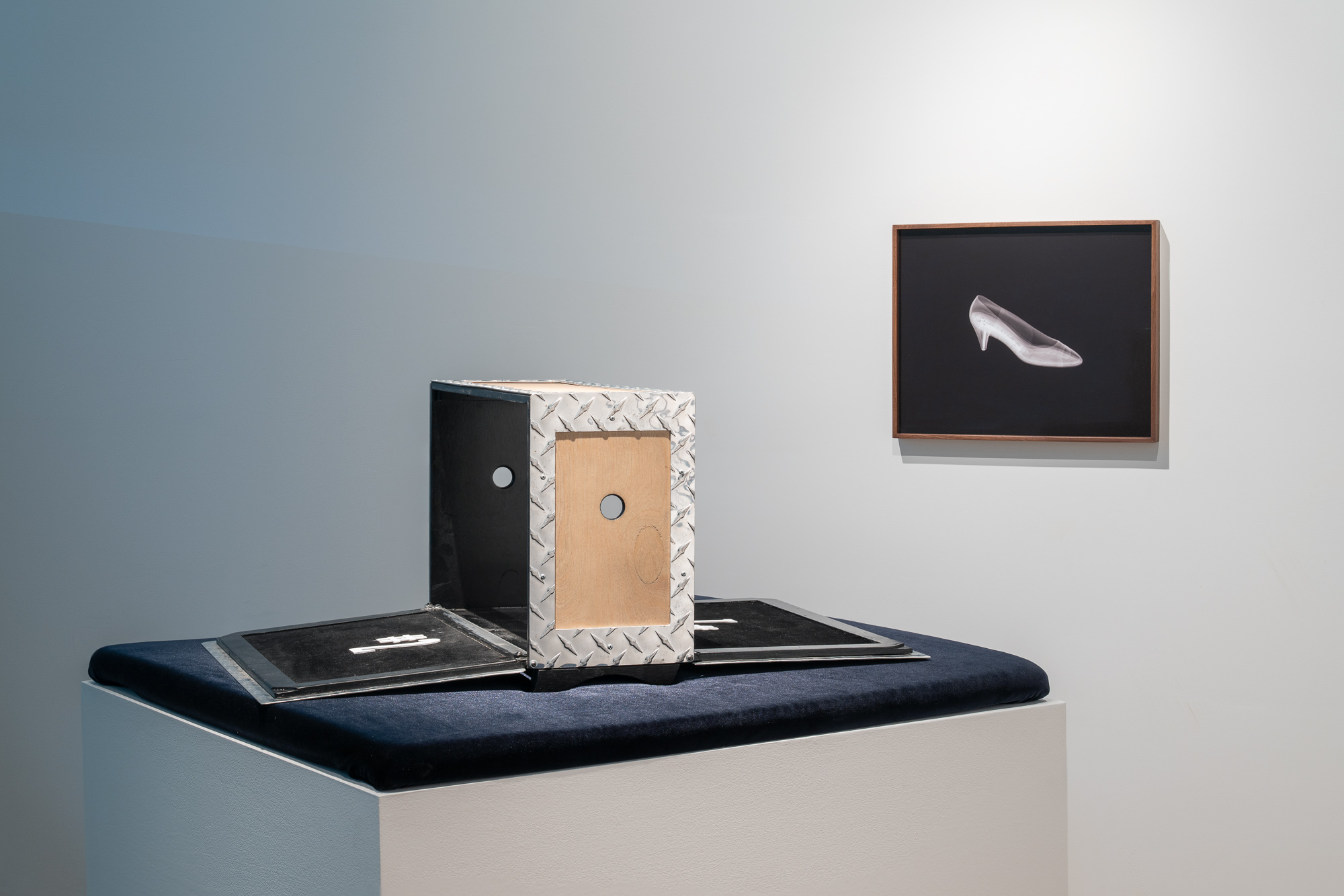 On a midnight blue velvet cushion on a white plinth is a wood and metal box with long sides opened and flapped down; on the wall an X-ray of a woman's low-heeled pump in a walnut frame
