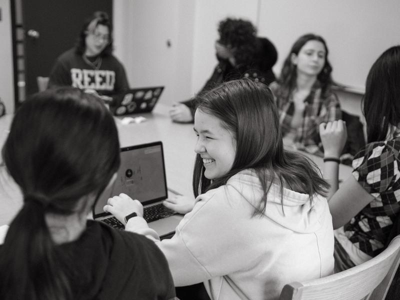 A group of students sit around a classroom table. One student on a laptop turns and smiles at another student.