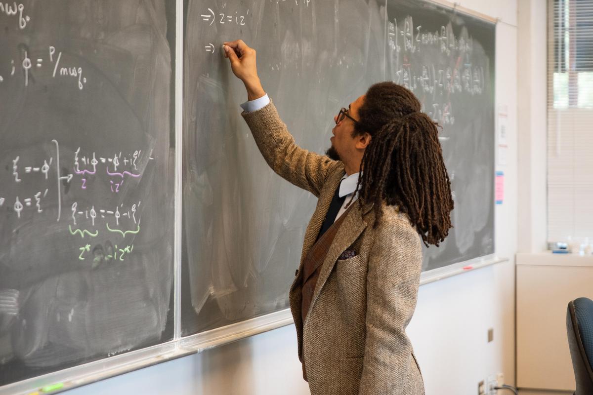 A professor writes an equation on a chalkboard while teaching a physics class.