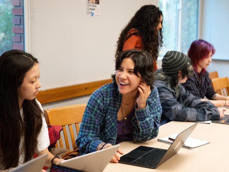 A student in a classroom leaning over an open laptop and smiling at another student.