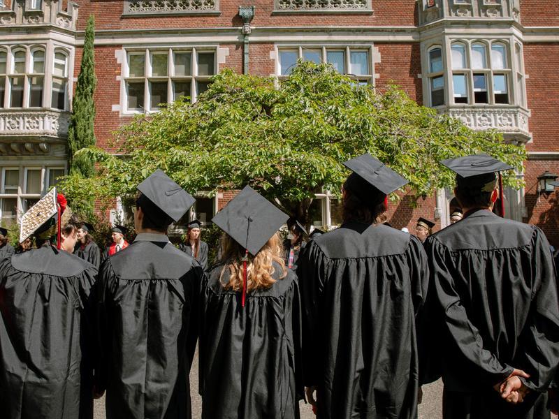 A row of Reed College graduates in academic regalia with their backs turned to the camera, looking up at Eliot Hall.
