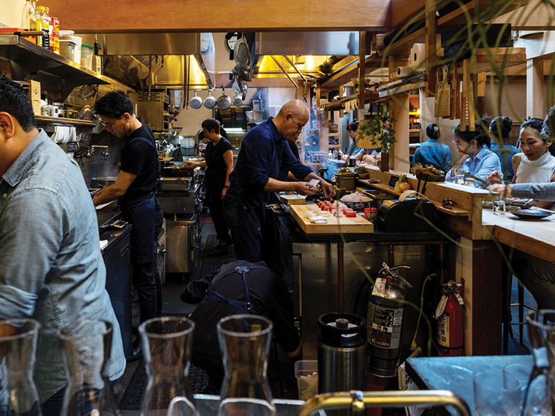 A lively restaurant kitchen with several chefs preparing food in front of customers sitting at a bar.