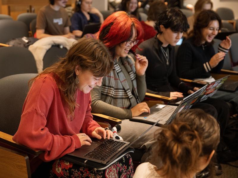 Students sit in rows in a lecture hall, looking at their computers and smiling.